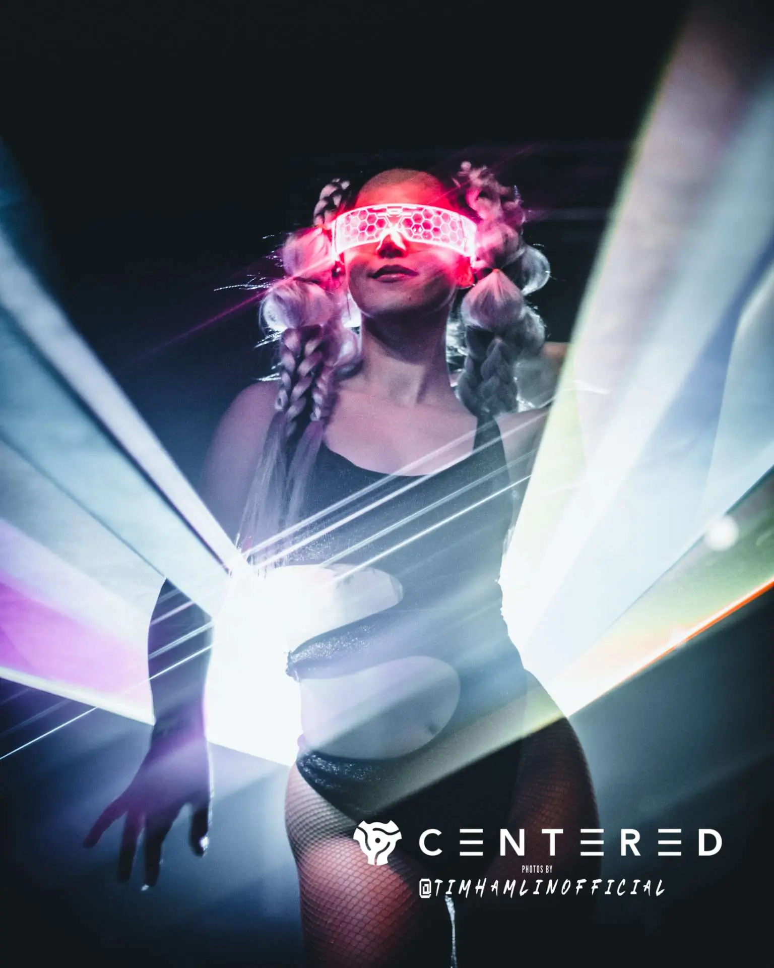 A young woman with blonde hair dances in a black leotard. She is illuminated by a bright colorful light from behind. She's wearing a bright neon lighted face sheild which gives her a robotic look.