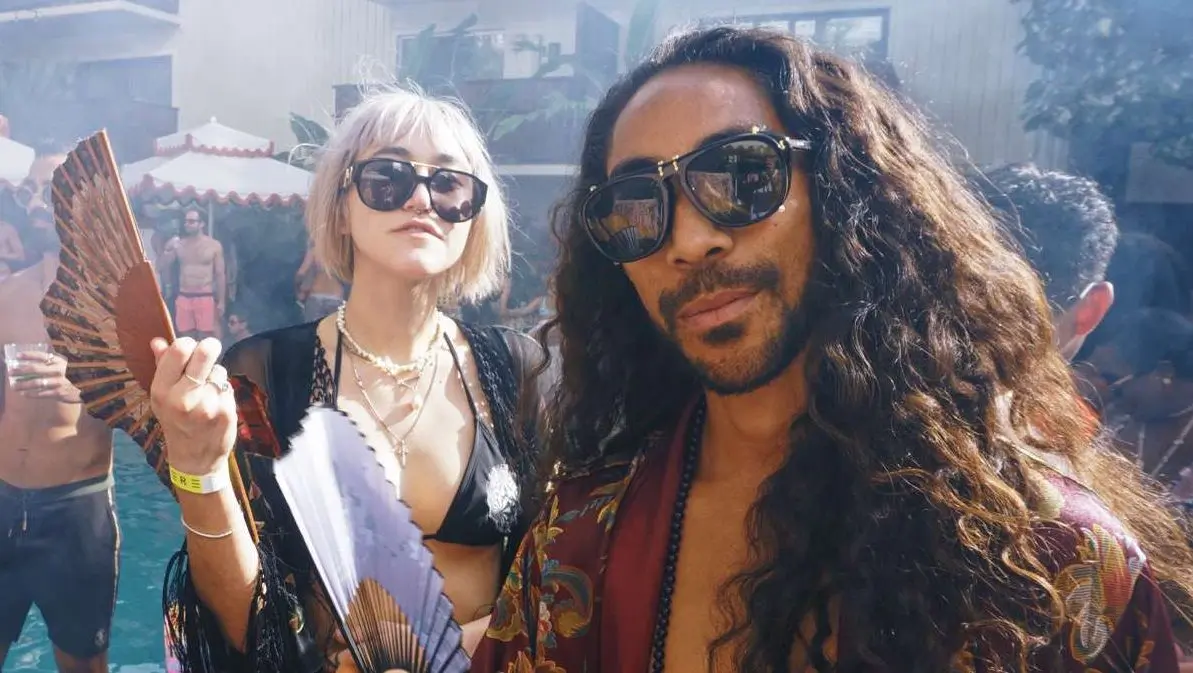 A man with dark long flowing hair wearing dark sunglasses and a silk robe stands near a pool deck. A young woman with blonde hair stands near him wearing a bikini top, and lots of dangling jewelry. They both stare directly at the camera and are holding asian hand fans.